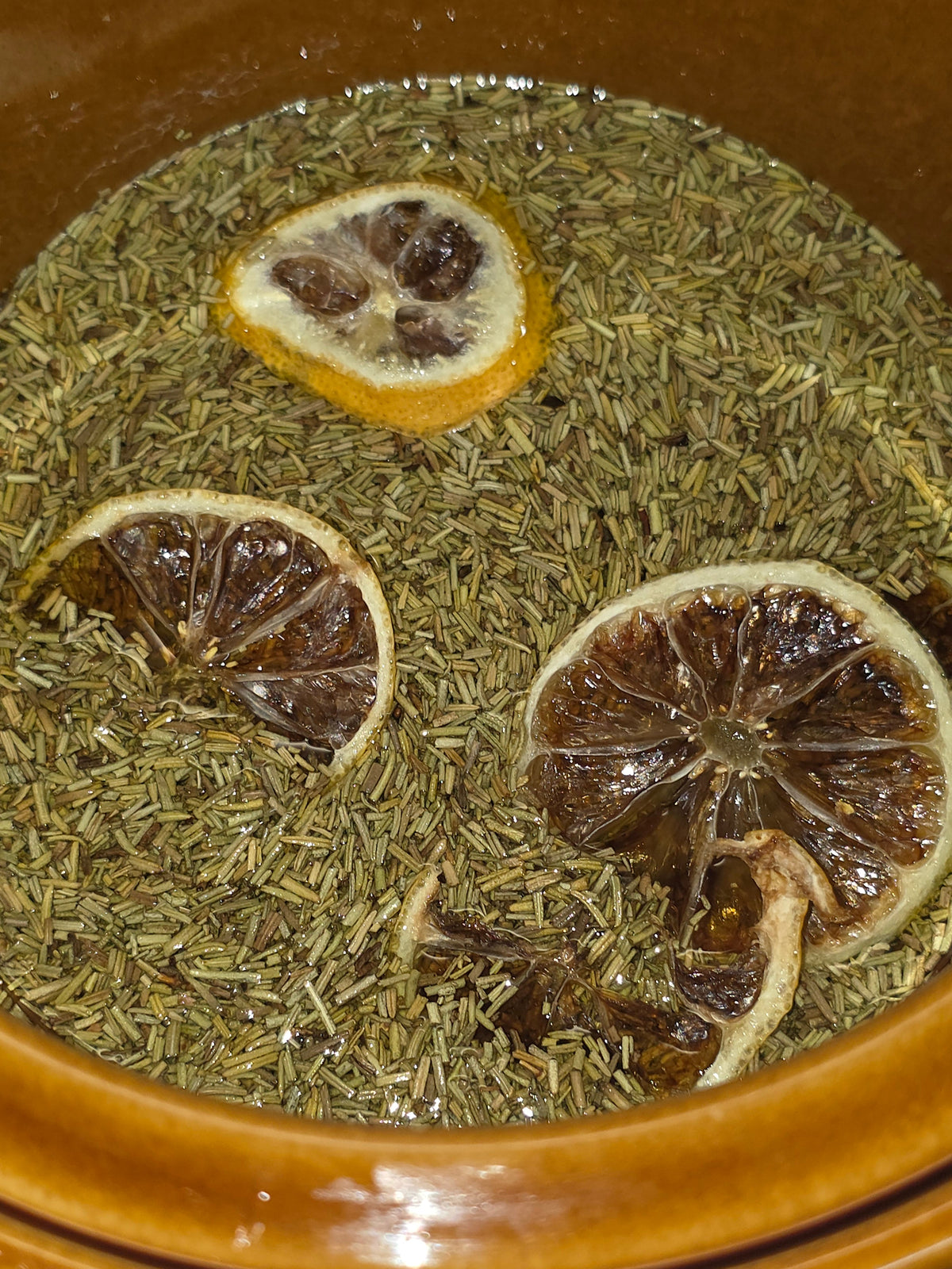 Overhead view of golden melted tallow in a copper pot topped with dried rosemary and dehydrated lemon slices during a slow botanical infusion